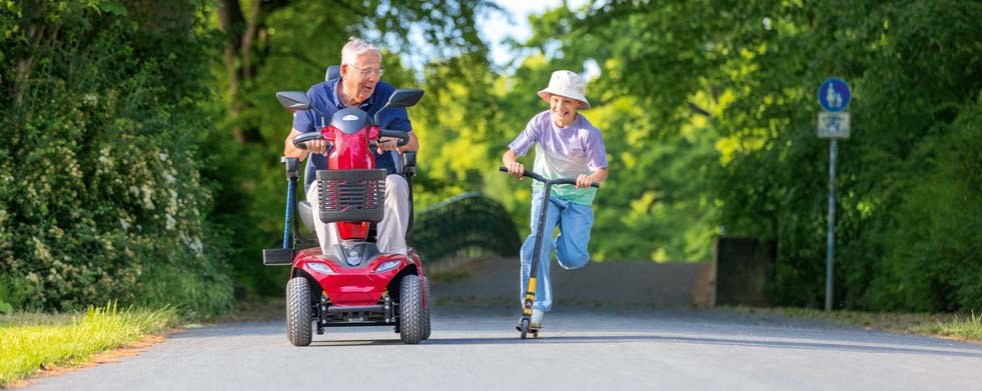 Fotografie zeigt älteren Mann auf rotem Elektromobil und Kind auf Tretroller, die nebeneinander auf einem von Bäumen gesäumten Weg ein Wettrennen fahren.