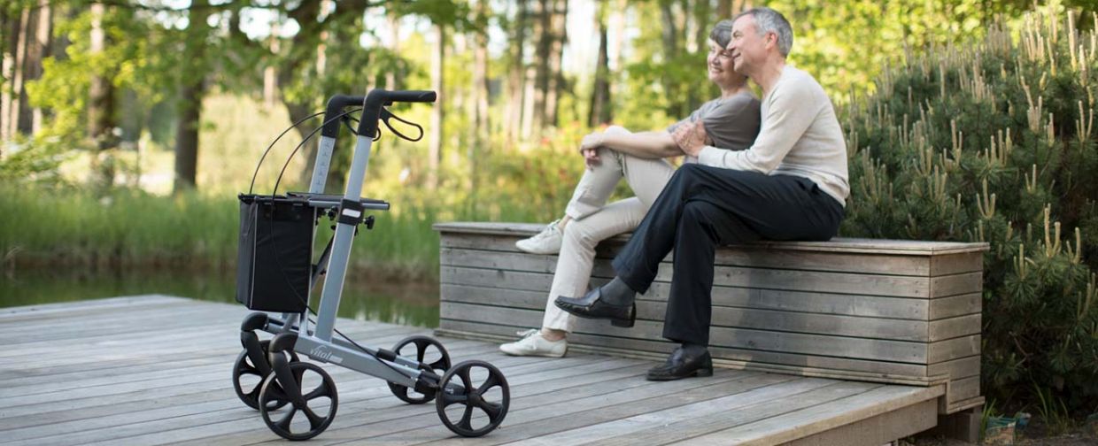 Ein Paar sitzt auf einer Bank an einem Holzpodest an einem Waldsee, davor steht ein RUSSKA Rollator vital plus