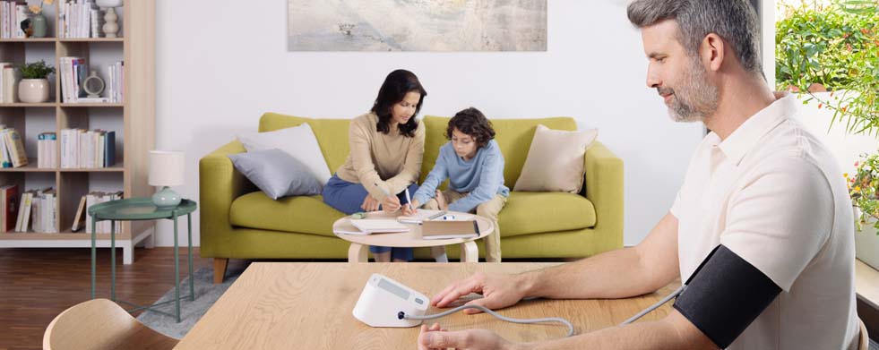 Ein Mann misst seinen Blutdruck mit dem OMRON Oberarm-Blutdruckmessgerät M2 Comfort. Auf einem Sofa im Hintergrund sitzen seine Frau und sein Kind. Das Kind schreibt etwas auf einen Block, die Mutter hilft ihm dabei. Die Atmosphäre ist entspannt.